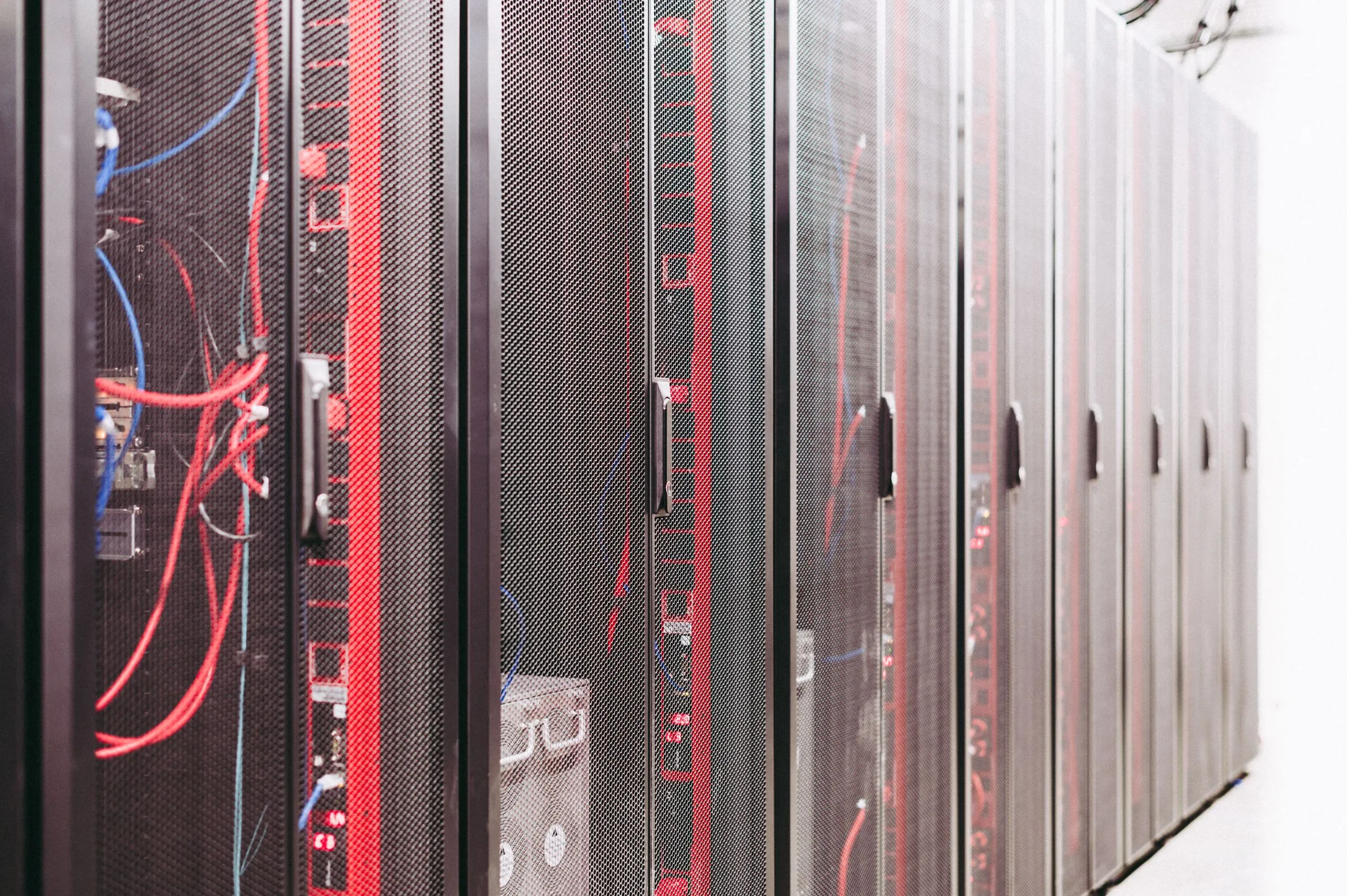A row of black server rack cabinets with perforated mesh doors in a white-walled data hall, red and blue patch cables visible through the open front of the nearest cabinet and silver door handles receding down the aisle