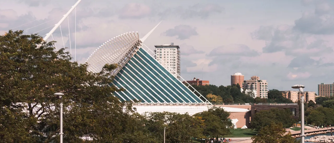 The Milwaukee Art Museum's white Calatrava wings and glass-and-steel pavilion seen from the lakefront in summer, with mature green trees and lamp posts in the foreground, a stone breakwater and the blue water of Lake Michigan at lower right, and mid-rise apartment towers along the bluff under a cloudy sky