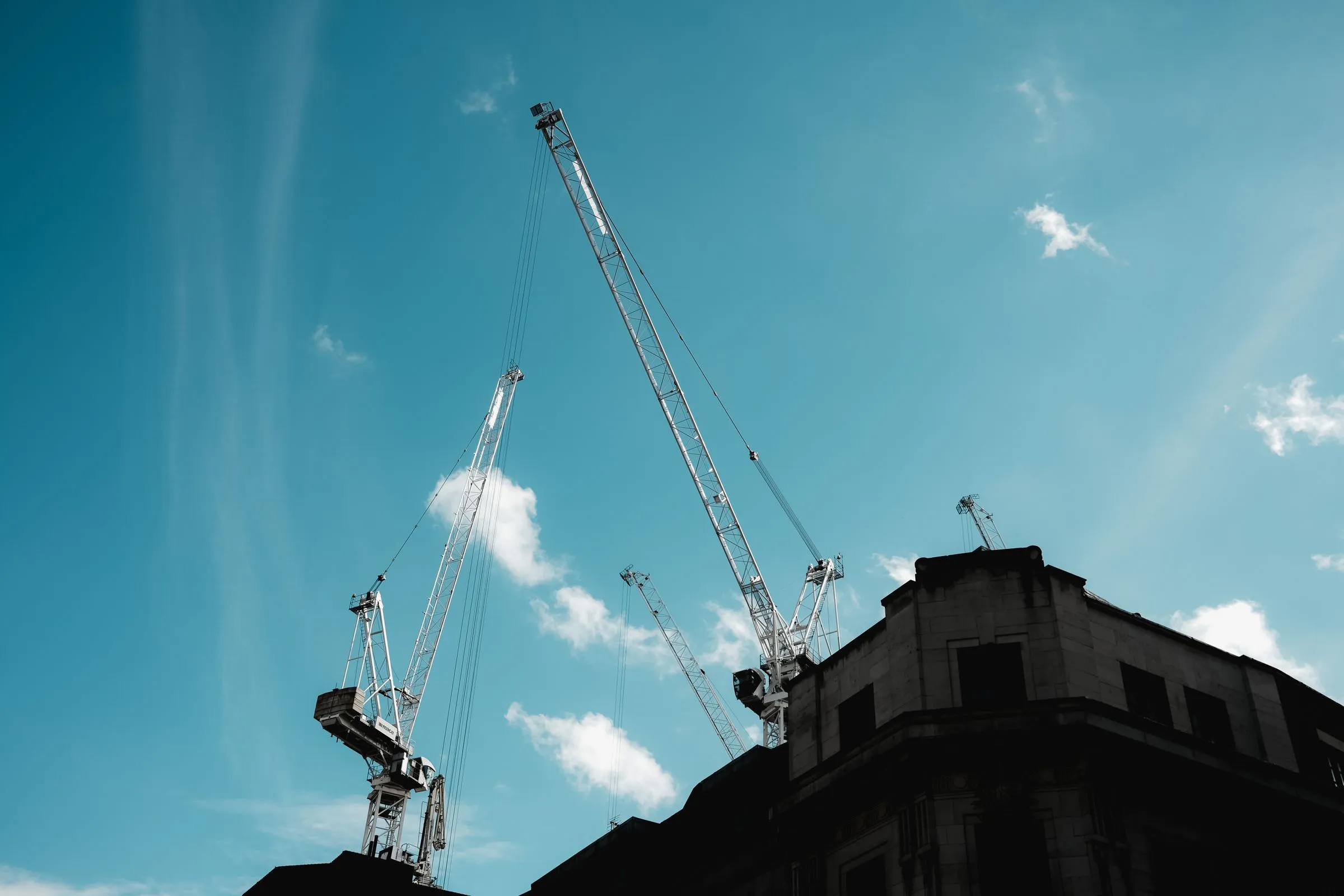 Tall construction cranes rising into a blue sky above the dark silhouette of an unfinished building
