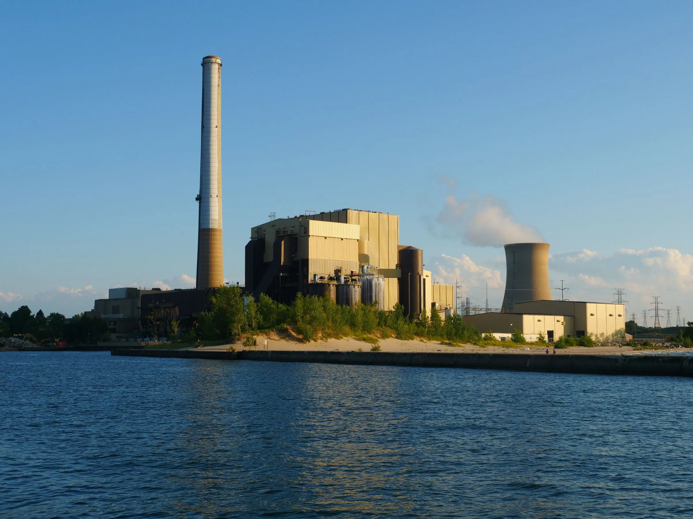 A power station with a tall smokestack and a single cooling tower emitting steam, standing on a sandy beach beside the deep blue water of Lake Michigan at dusk
