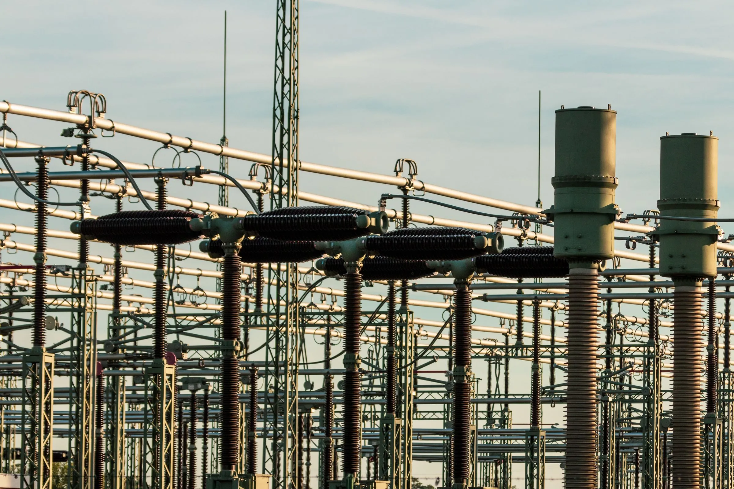 Rows of high-voltage insulators, disconnect switches, and aluminium bus tubing in an outdoor transmission switchyard, lit by low evening sun