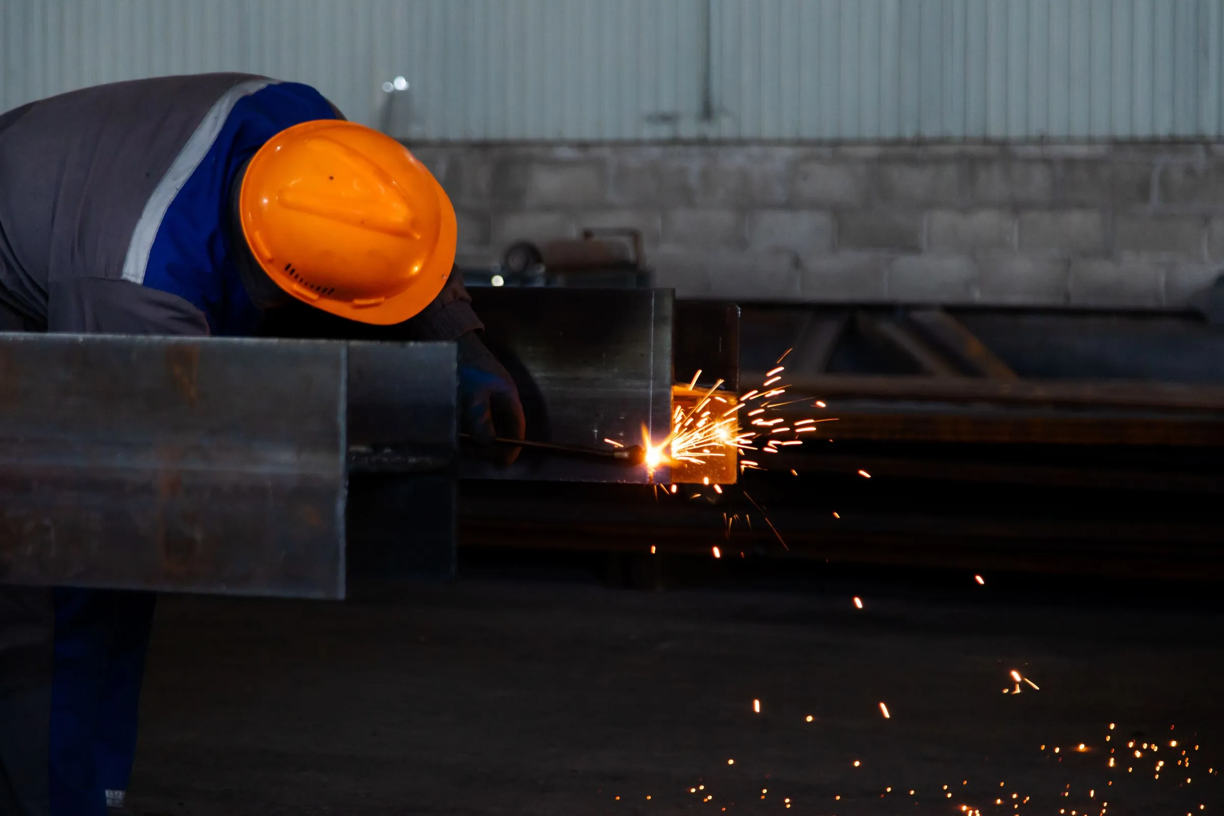 A worker in an orange hard hat bending over a heavy steel box section, cutting it with a torch as orange sparks scatter across a dark fabrication shop floor