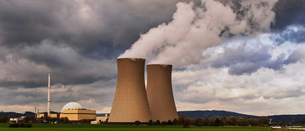Two concrete cooling towers of a nuclear power plant releasing plumes of steam under a dark overcast sky, with a domed reactor building and green fields in the foreground