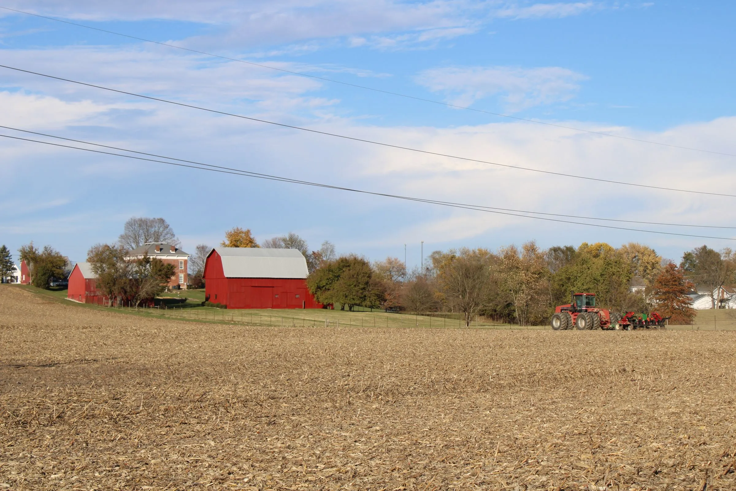 An Ohio farm in late autumn with a red barn, a brick farmhouse, and a tractor at the edge of a harvested cornfield, with transmission lines crossing the sky overhead