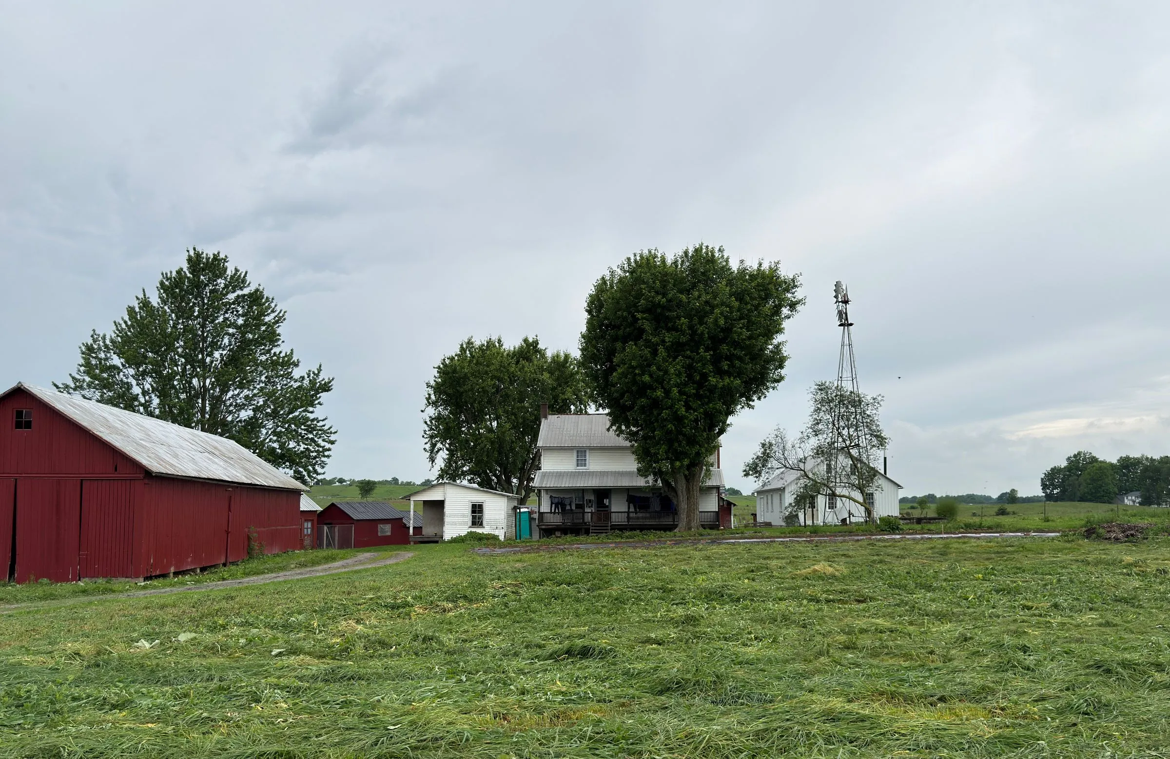 A farm in rural Ohio under an overcast sky, with a weathered red barn, a white farmhouse shaded by a large tree, a windmill, and a freshly cut hay field in the foreground