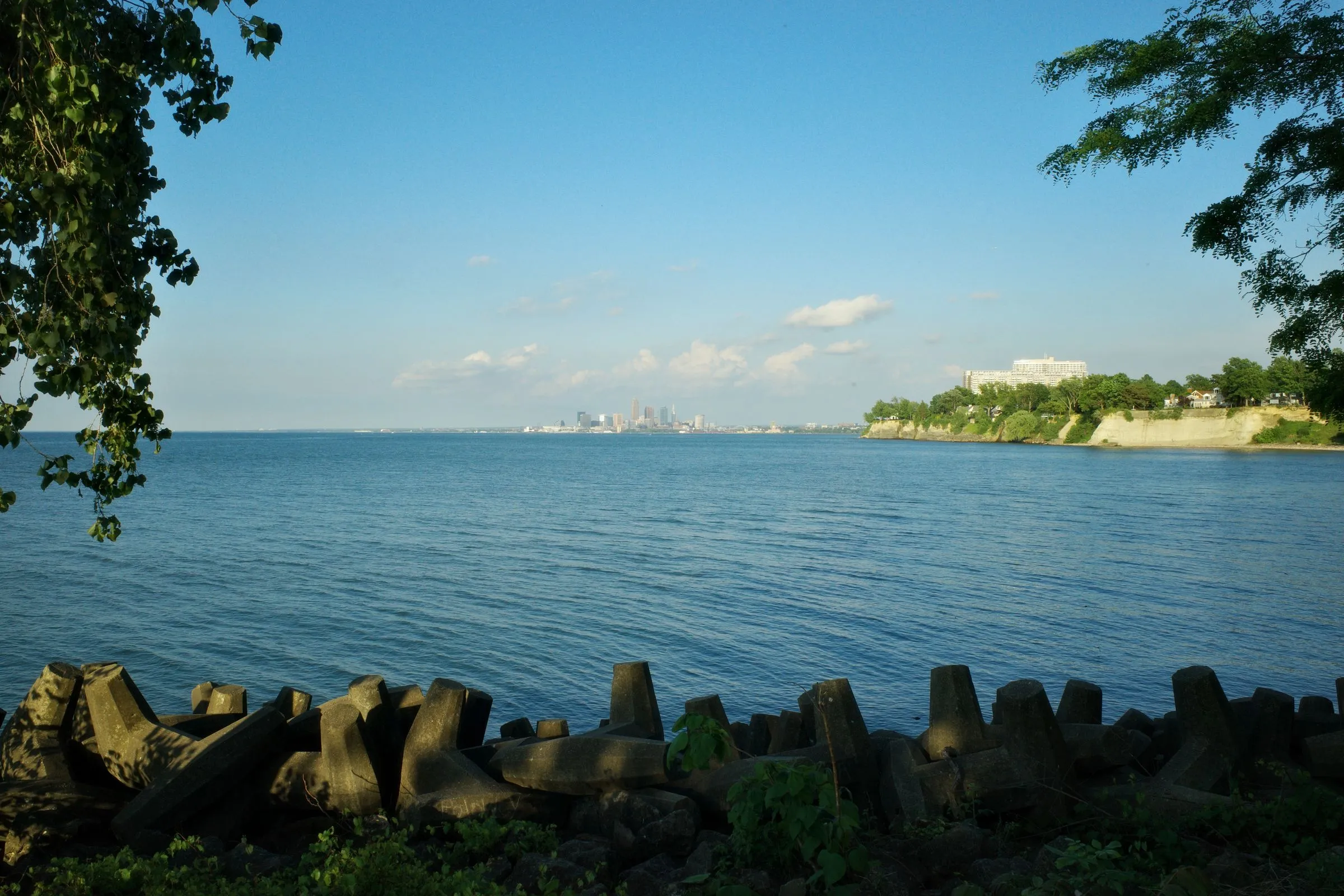 The downtown Cleveland skyline seen across the calm blue water of Lake Erie from a lakeside park, framed by trees, with a stone breakwater in the foreground