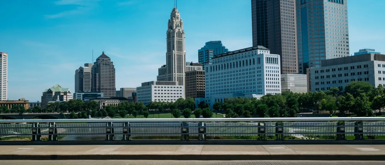 The downtown Columbus, Ohio skyline on a clear summer day seen from a bridge over the Scioto River, with the LeVeque Tower's stepped spire in the center and the Rhodes State Office Tower at right
