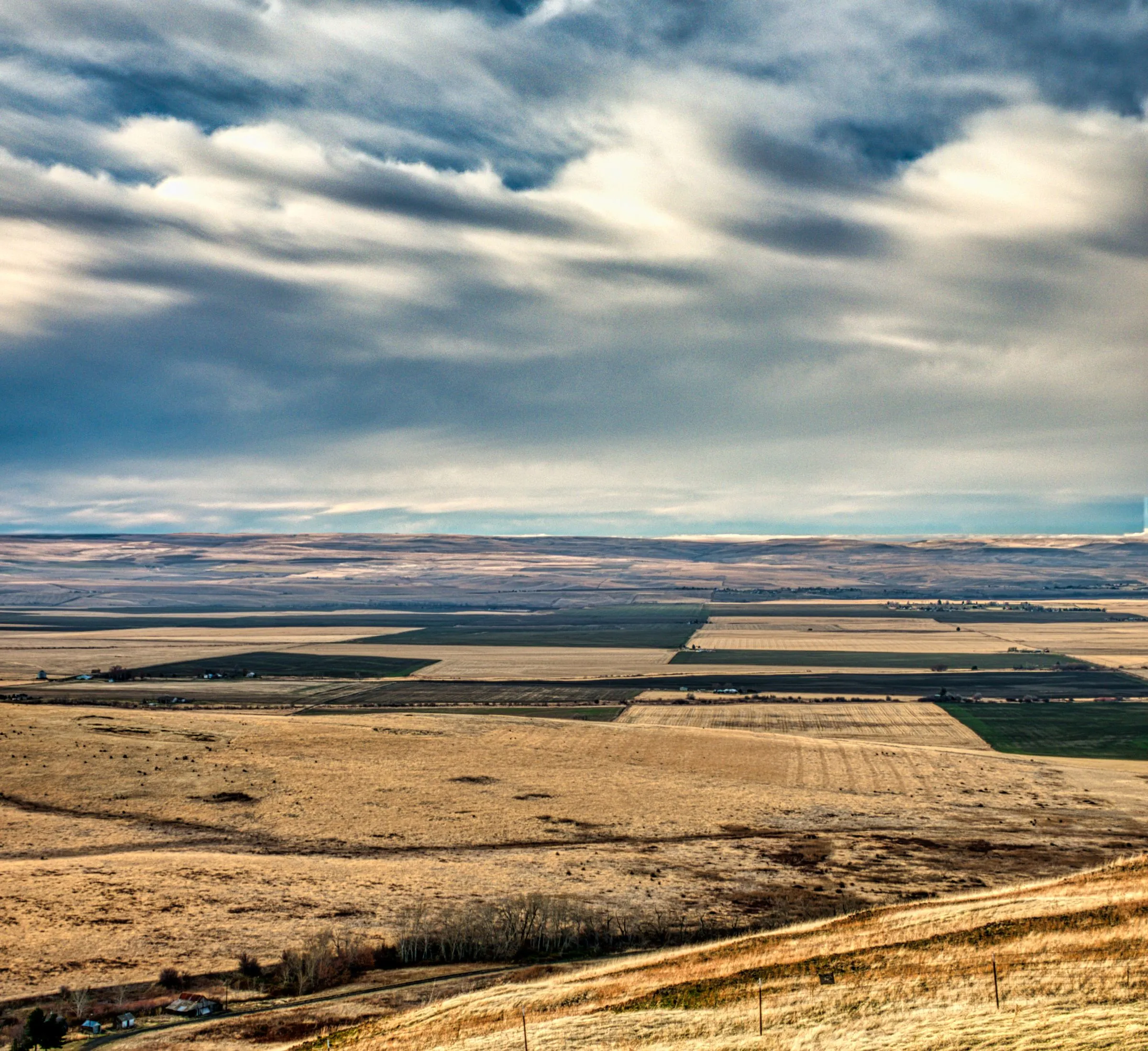 Aerial view across the Umatilla County farm country near Pendleton, Oregon, with tan dryland wheat stubble and green irrigated fields laid out in rectangles on a broad plateau, a dirt road and a few farm buildings in the foreground, low brown hills on the horizon, and a bank of gray and white cloud streaking across the sky