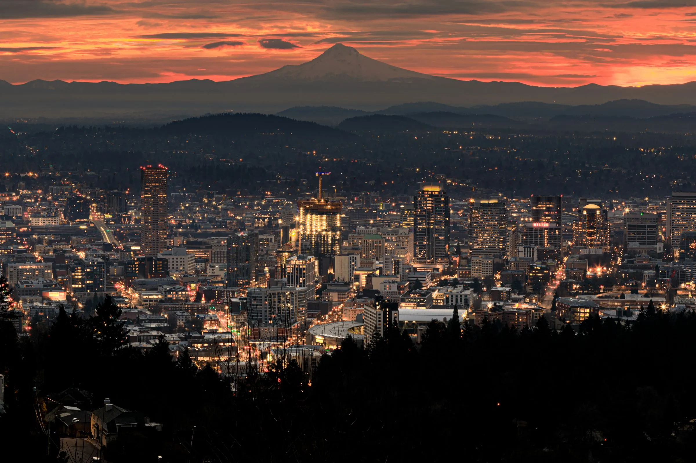 The downtown Portland, Oregon skyline at dusk seen from the West Hills, with office towers lit against a dark grid of streets, the forested ridges of the east side behind them, and the snow-covered peak of Mount Hood on the horizon under an orange and red sky