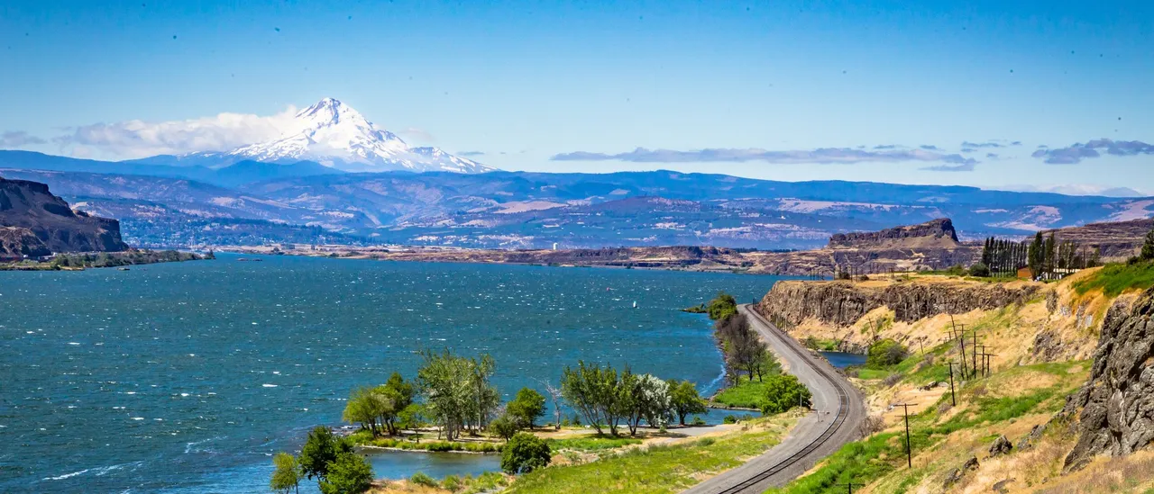 The Columbia River east of The Dalles, Oregon, on a windy summer day, with whitecaps on the deep blue water, a railroad track curving along the grassy shore in the foreground, basalt cliffs on the far bank, and the snow-covered cone of Mount Hood rising above a band of cloud under a clear sky