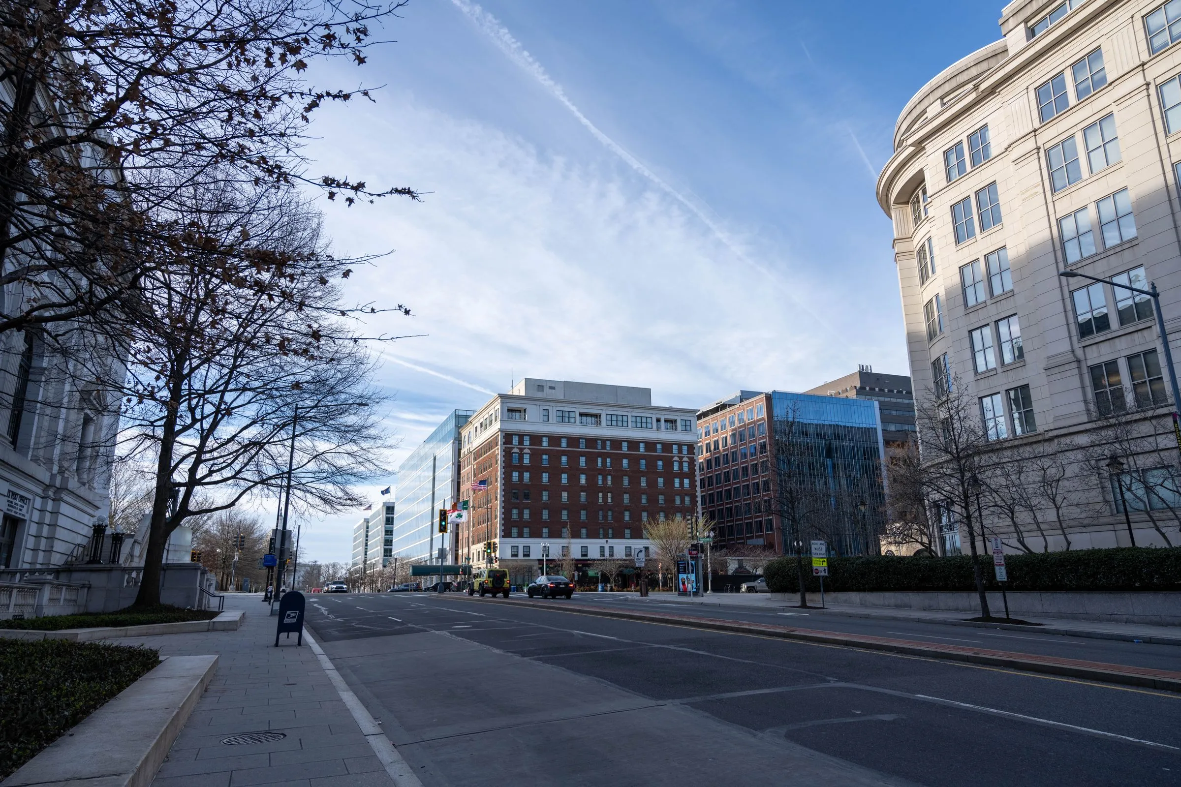 A quiet downtown Washington, D.C. street in winter, with a bare tree and a stone federal-style building on the left, a blue mailbox on the sidewalk, and a row of brick and glass office buildings with American flags under a blue sky
