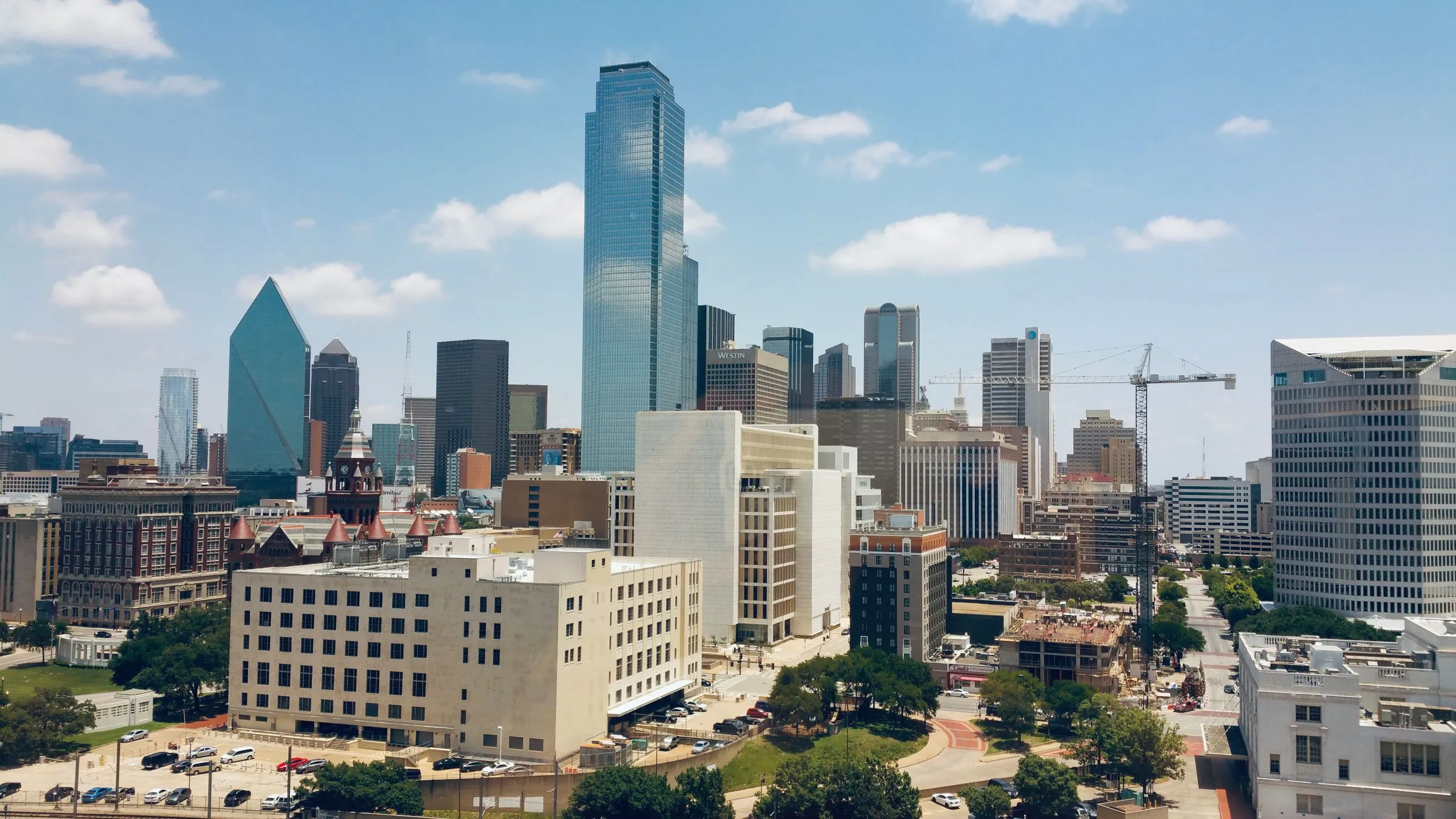 Aerial view of downtown Dallas under a clear blue sky, with Bank of America Plaza, Fountain Place, and Reunion Tower rising above lower office blocks and a construction crane