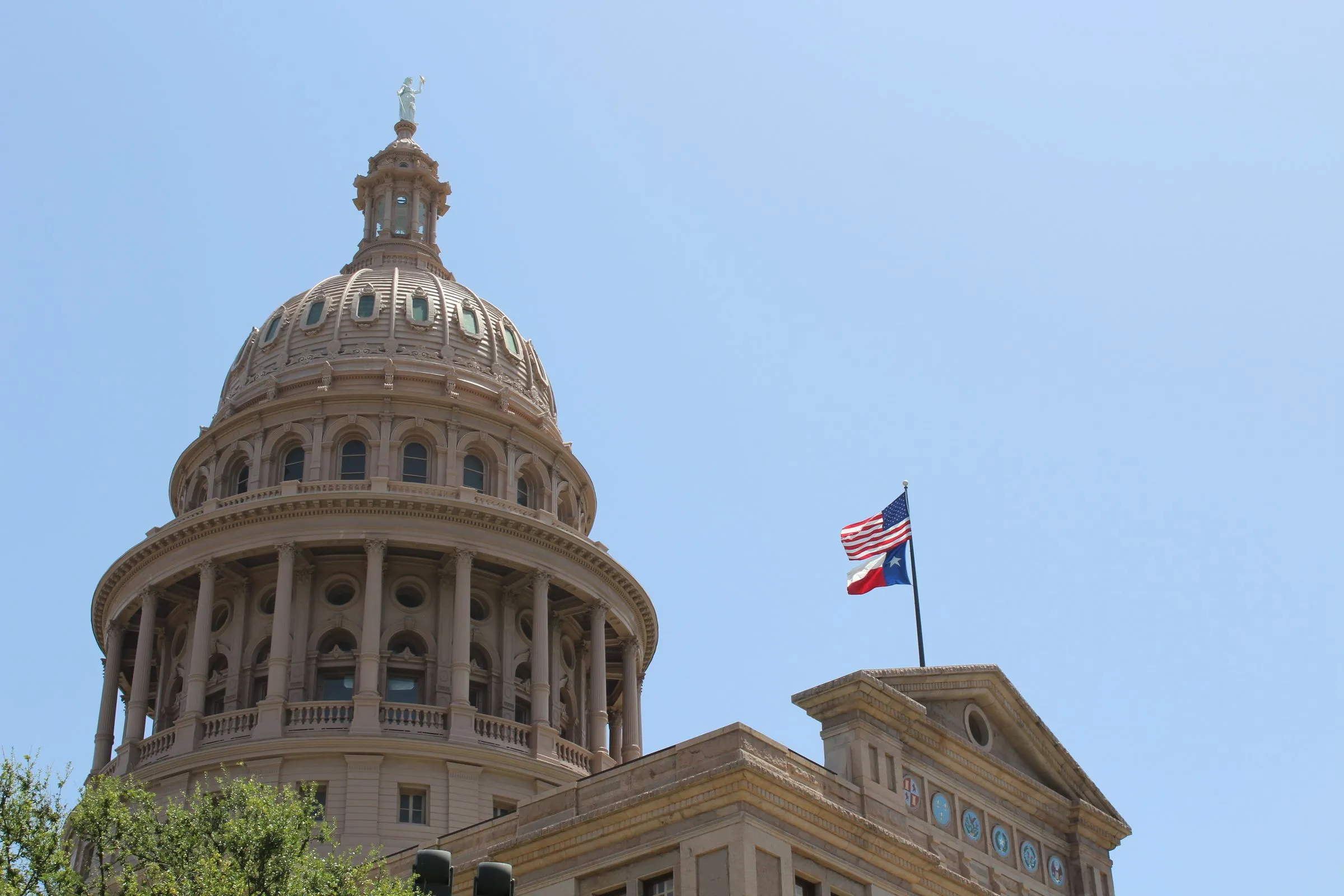 The dome of the Texas State Capitol in Austin against a clear sky, with the United States and Texas flags flying from the roof below it