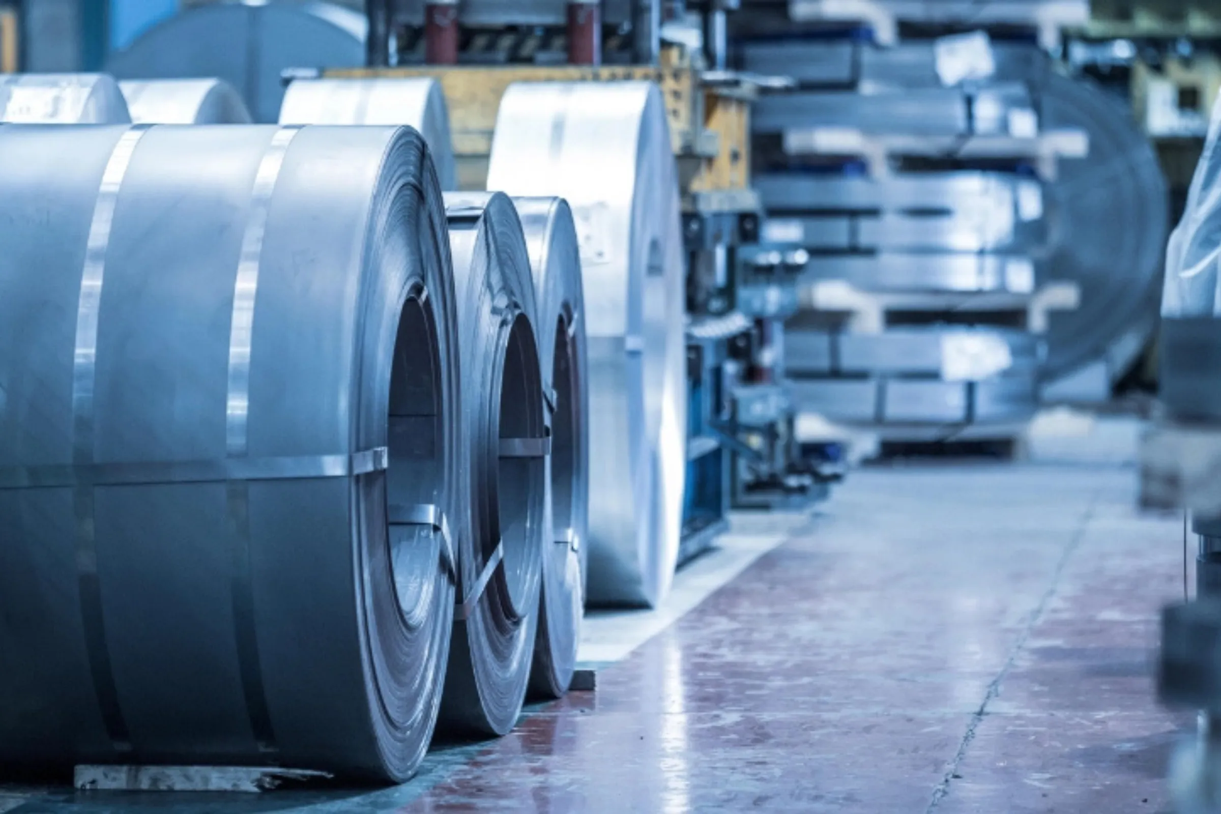 Rows of large rolled steel coils standing on a factory floor, with more coils stacked in the background of an industrial hall