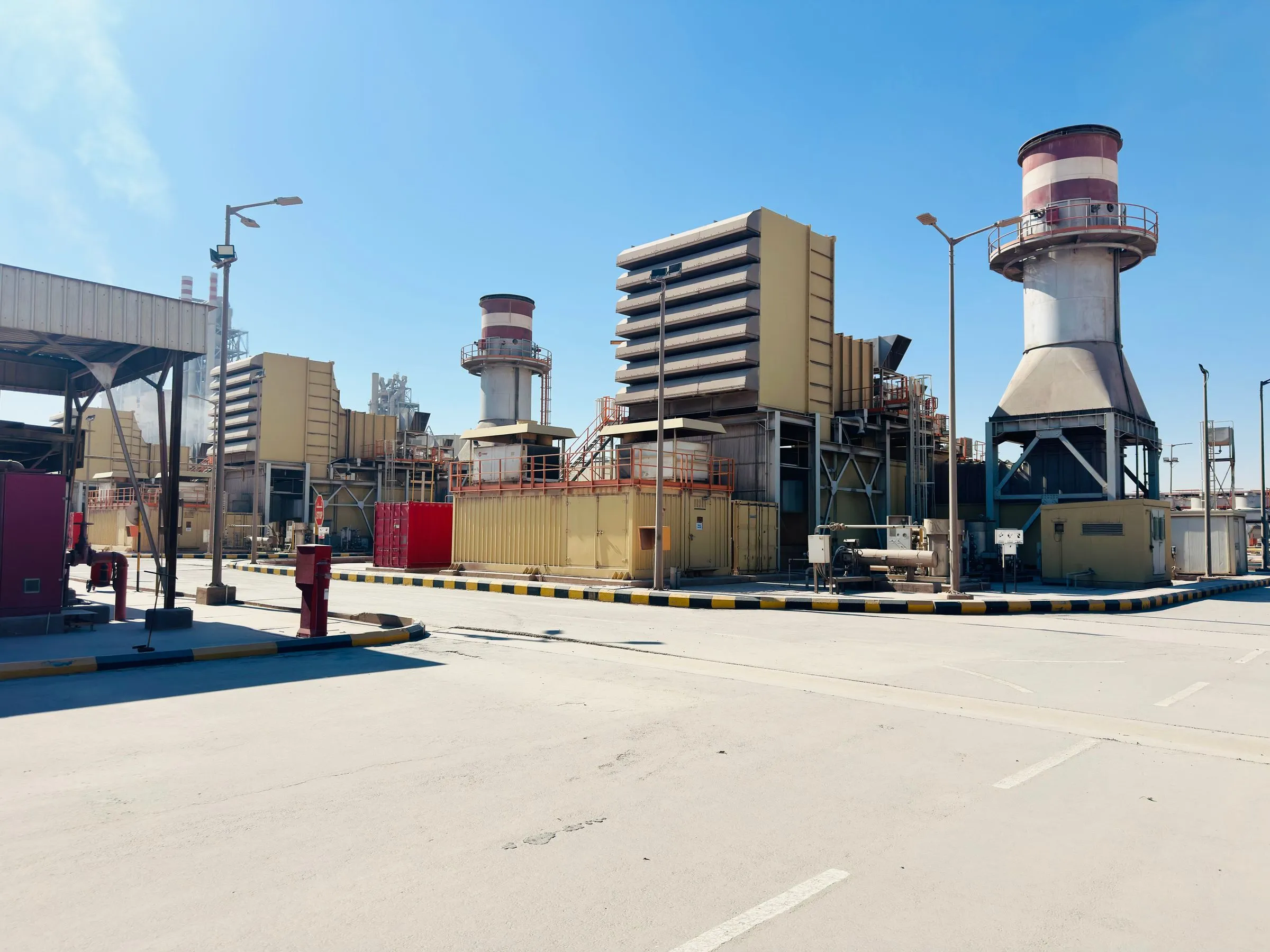 Gas turbine generating units at a power plant under a clear blue sky, with tall exhaust stacks and stacked air-intake filter houses beside a paved access road