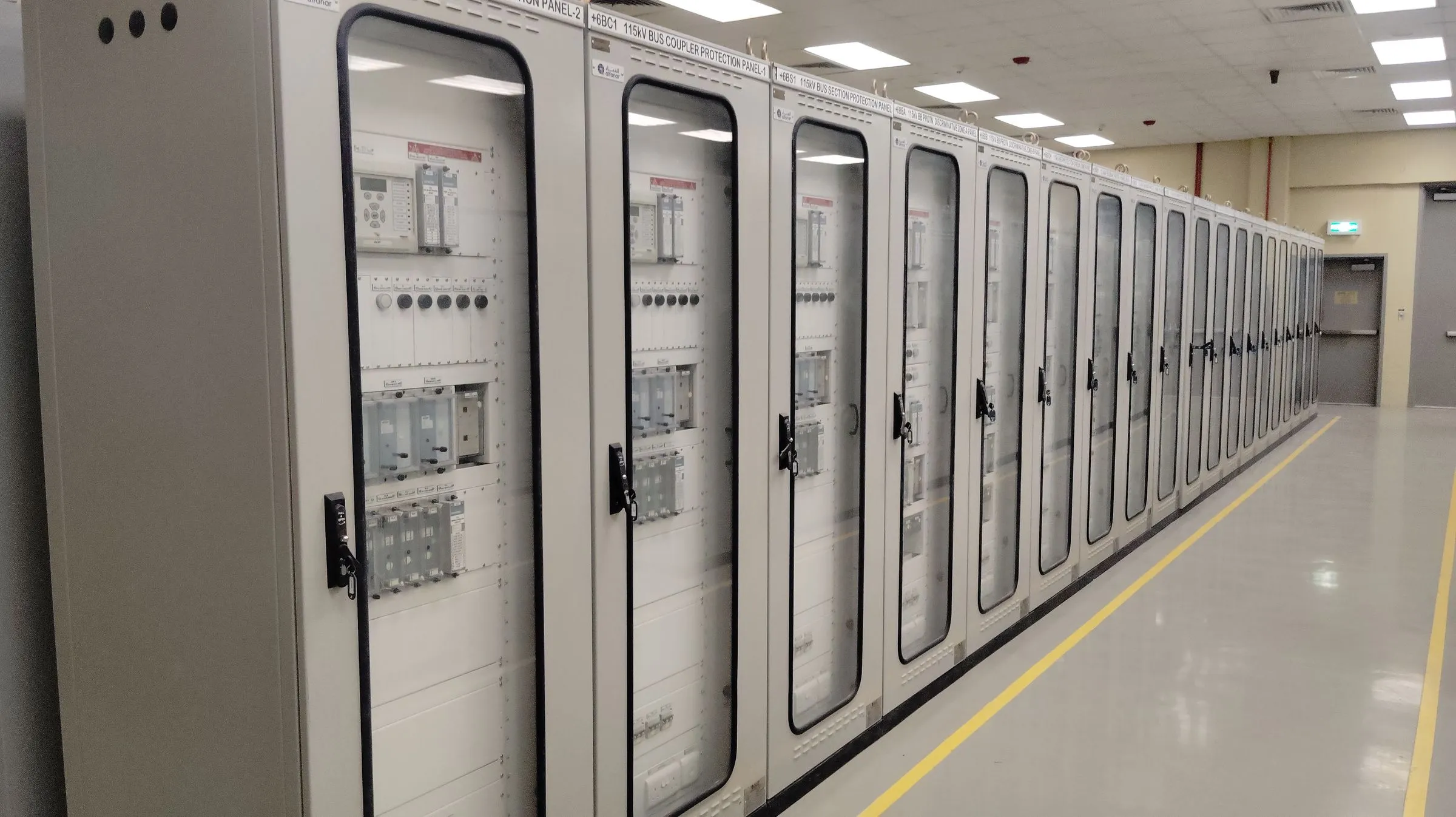 A row of tall grey protection and switchgear cabinets with glass doors lining a bright electrical control room