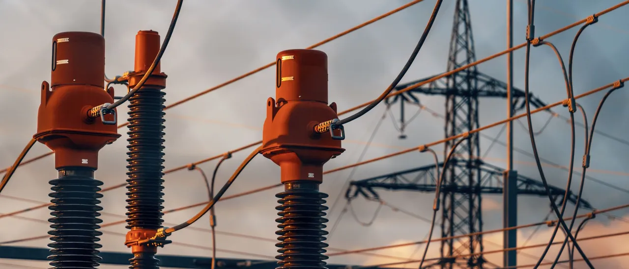 Three orange-topped high-voltage bushings on substation equipment, with a lattice transmission tower and power lines behind them at sunset