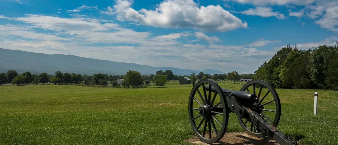 A Civil War cannon on the green lawn of Manassas National Battlefield Park in Prince William County, Virginia, with a treeline and the Bull Run Mountains under a blue sky with scattered clouds