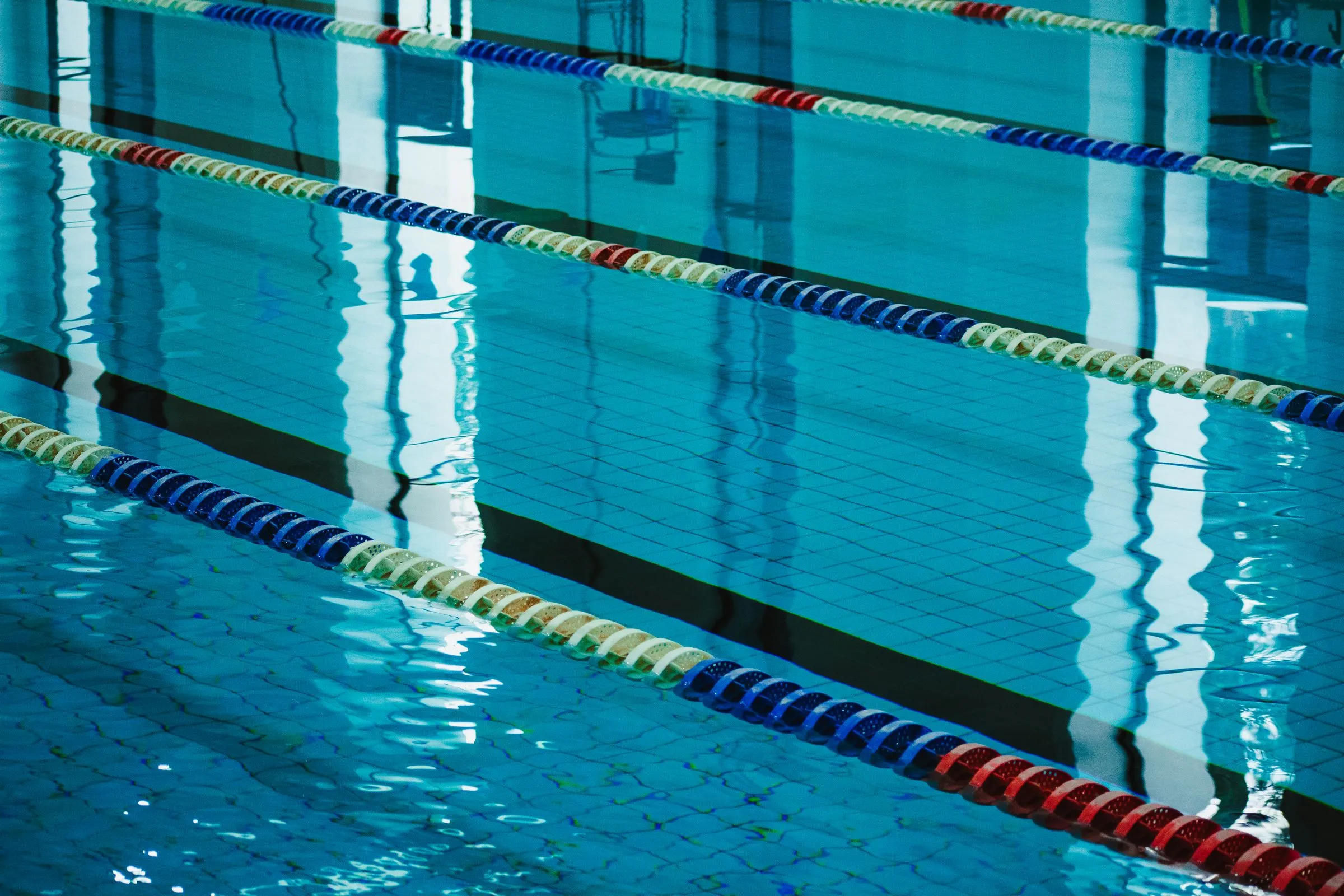 Empty indoor swimming pool with red, white, and blue lane divider ropes stretched across calm turquoise water