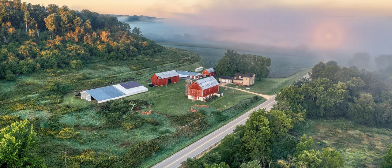 Aerial view of a Wisconsin dairy farm with red barns and a silo beside a two-lane road, with morning fog over the fields behind it