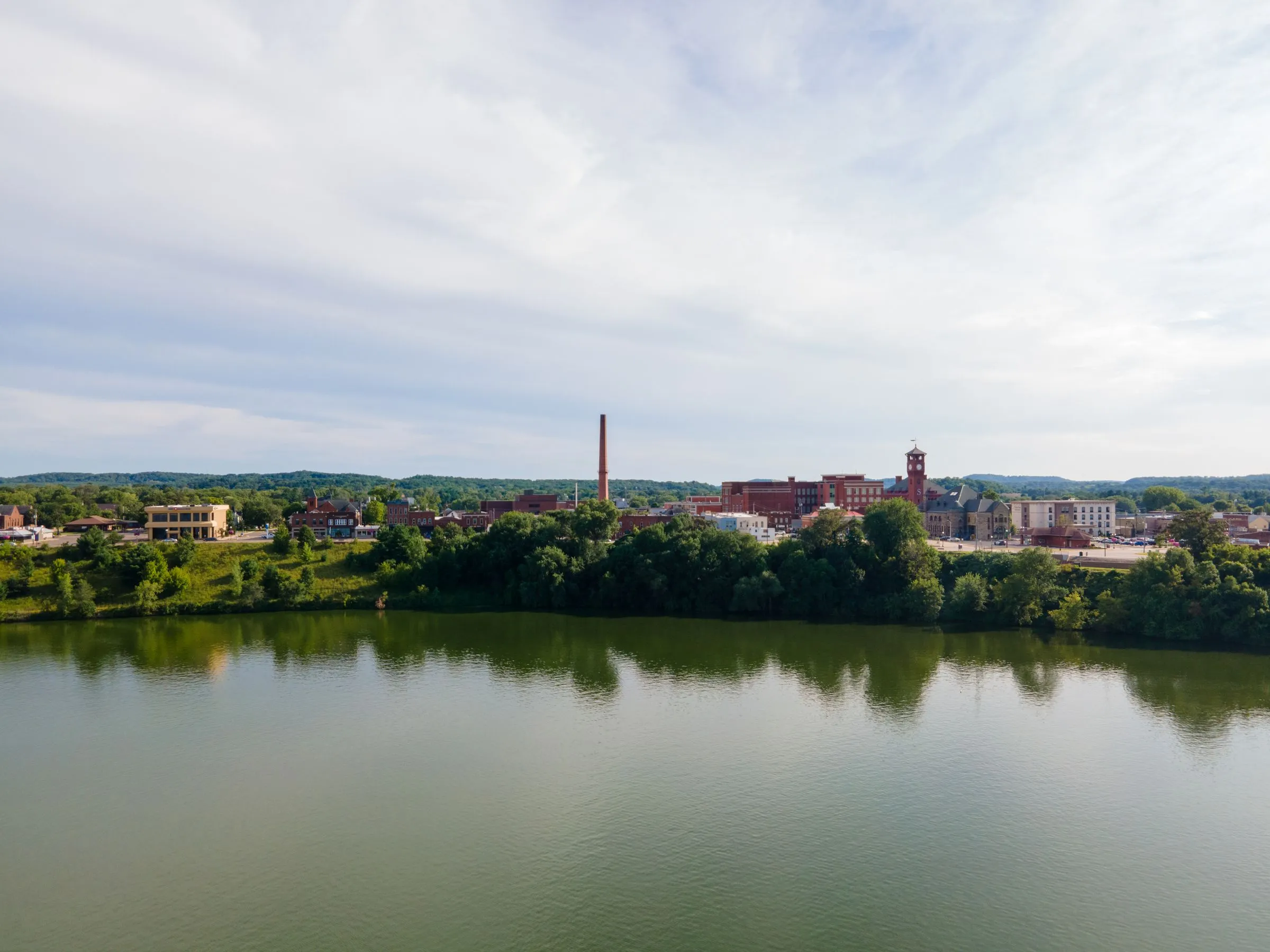 Aerial view of Menomonie, Wisconsin across Lake Menomin on the Red Cedar River, with the downtown buildings, a tall brick smokestack, and a church steeple rising behind the tree-lined shore