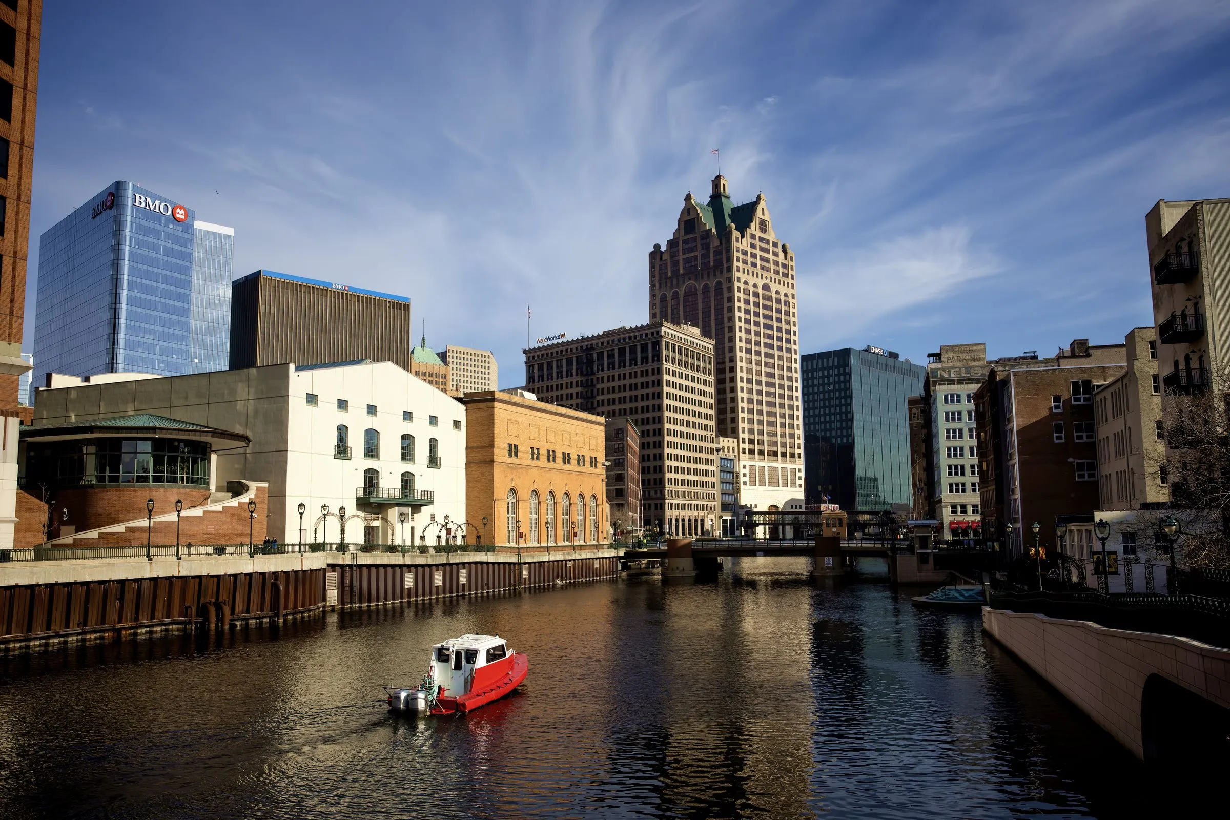 Downtown Milwaukee seen along the Milwaukee River, with a small red tour boat on the water, the ornate Milwaukee City Hall tower and office buildings behind, and a bridge crossing in the distance
