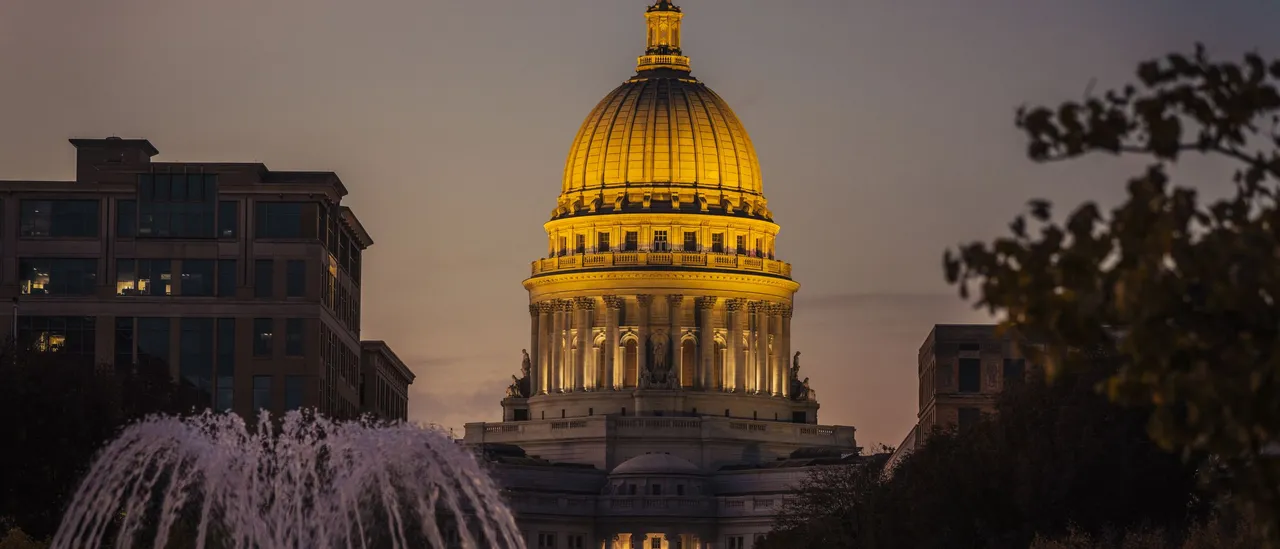 The gilded dome of the Wisconsin State Capitol in Madison lit gold at dusk, with a fountain spraying in the foreground and downtown office buildings to the left